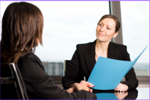 women holding a blue folder with smile looking at another women at table for interview. Back of head is showing on person being interviewed