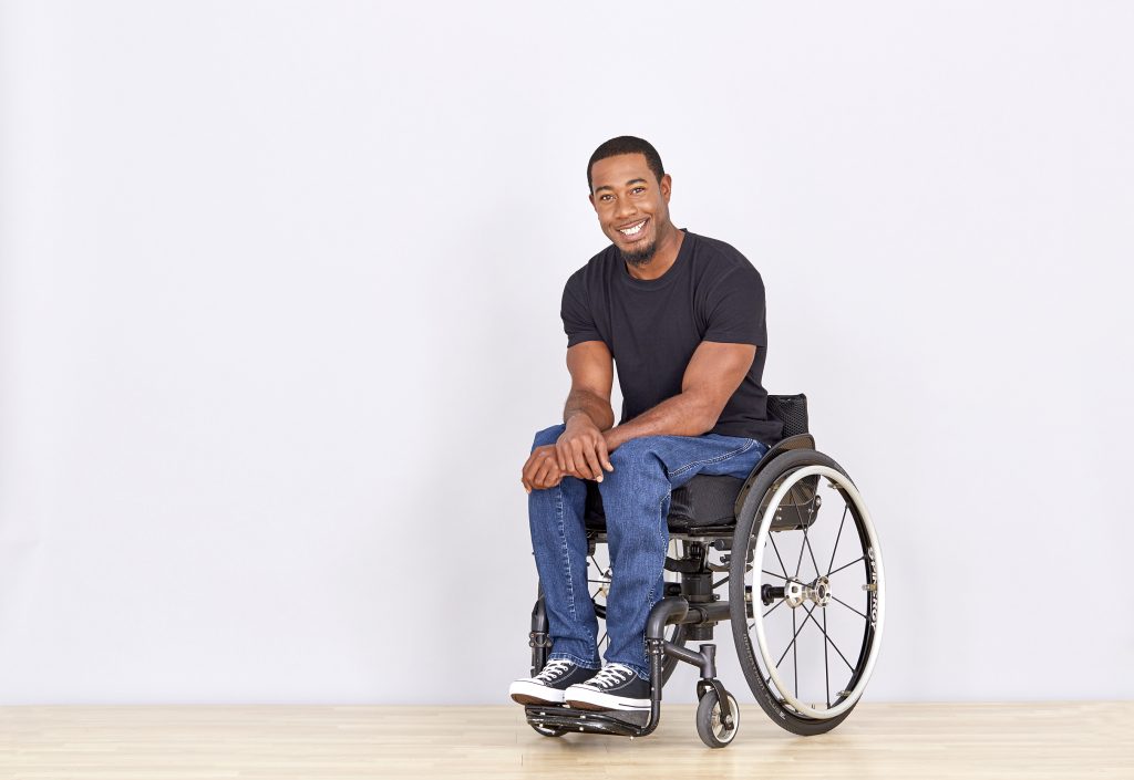 Anthony Lue smiles at the camera while seated in a wheelchair against a light background. He wears a black T-shirt, jeans and sneakers.