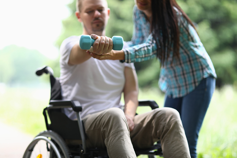 Female physiotherapist helps disabled man in wheelchair to lift weight of arm in park